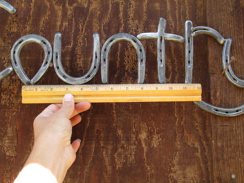 May include: The word "Country" spelled out using horseshoes, with a ruler measuring the length of the word. The horseshoes are silver in color and are nailed to a brown wooden surface.
