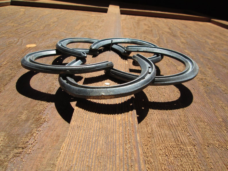 May include: Five silver horseshoes are interlocked to form a decorative design. The horseshoes are arranged in a circular pattern, with the points of the shoes facing outwards. The horseshoes are mounted on a wooden surface.