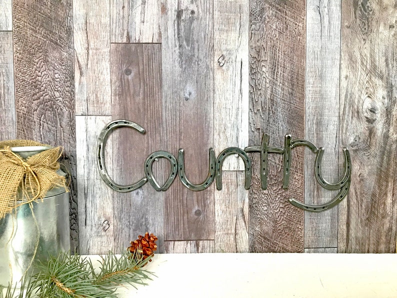 May include: The word "Country" spelled out using horseshoes. The horseshoes are silver in color and are mounted on a wood background.
