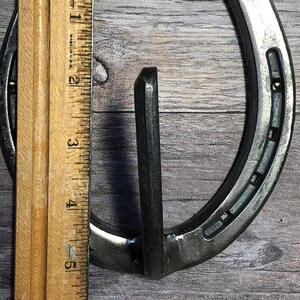 May include: A silver horseshoe with a metal hook attached to the top. The horseshoe is about 6 inches tall and is measured against a ruler.