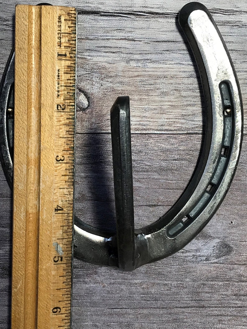 May include: A silver horseshoe with a metal hook attached to the top. The horseshoe is about 6 inches tall and is measured against a ruler.