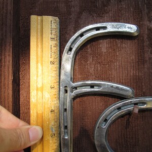 K&ouml;nnte beinhalten: Zwei silberne Hufeisen sind miteinander verbunden, um den Buchstaben "F" zu bilden. Die Hufeisen sind etwa 15 cm hoch und werden an einem Holzlineal gemessen.