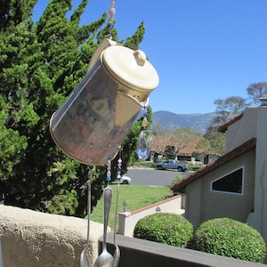May include: A vintage-style coffee pot wind chime with a floral pattern and three silver spoons hanging from it. The wind chime is made of metal and has a beaded chain with colorful beads.