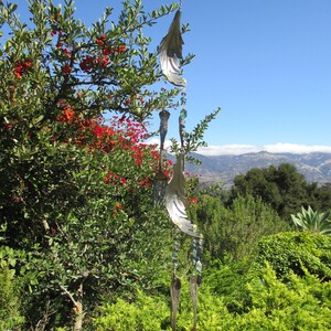 May include: A handcrafted wind chime with a unique design, featuring repurposed silverware and decorative metal wings. The chime hangs from a string of colorful beads and is set against a backdrop of lush green foliage and a clear blue sky.