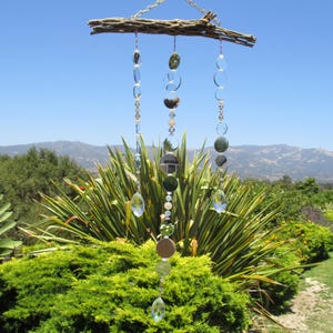 May include: A hanging suncatcher featuring a piece of weathered driftwood at the top, from which three vertical strands of clear glass crystals and circular reflective mirror discs are suspended, creating a decorative mobile with light-catching elements.