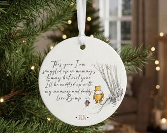 May include: A round white ceramic Christmas ornament hanging from a white ribbon. It features a black ink illustration of Winnie the Pooh and Piglet, with the text: This year I am snuggled up in mummy's tummy but next year I'll be cuddled up with my mummy and daddy. love Bump. 2026.