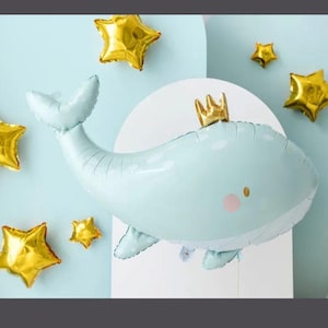 Può includere: Un palloncino a forma di balena azzurra con una corona d'oro e guance rosa. Intorno, diversi palloncini a forma di stella dorata, il tutto su uno sfondo azzurro. Perfetto per un baby shower o una festa di compleanno.