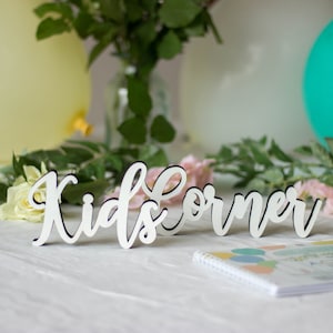 May include: White wooden sign with the words "Kids Corner" in cursive script. The sign is on a white table with a notebook and flowers in the background.