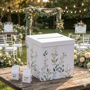 May include: A white wedding card box with a slot on top, decorated with floral patterns in pink, green, and beige. Two small white favor boxes with heart-shaped cutouts sit in front of the card box. The background shows a wedding ceremony setting.