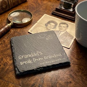 May include: A square slate coaster with rough, natural edges featuring an engraved graphic of a teacup and cookies. The text reads: Grandad's Break from Grandma :) Love Henry and Gracie rock. The dark gray stone surface has a textured finish.