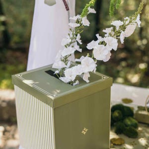 May include: A square olive green card box featuring a white vertical striped pattern on the sides and a thin white border on the lid. A narrow rectangular slot is centred on the top, containing several white artificial flowers with green stems.