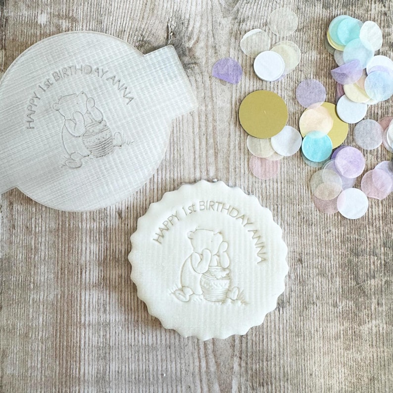 May include: A white, round cookie with a scalloped edge, featuring a Winnie the Pooh design and the text "HAPPY 1ST BIRTHDAY ANNA." A matching, irregularly shaped item with the same design is next to the cookie. Colorful confetti is scattered around the items.