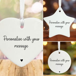 May include: Three white ceramic hanging ornaments: a heart, a star, and a circle. Each has a white ribbon and the text "Personalise with your message". The heart and circle ornaments have a small black heart.
