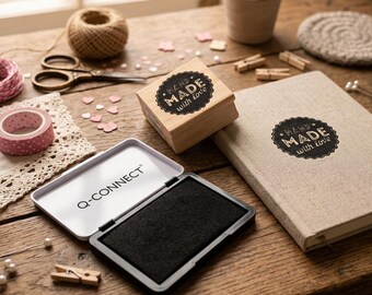 May include: A wooden stamp with a black circular label reading HAND MADE with love, a beige notebook with the same label, and an open black Q-CONNECT ink pad. Also includes a ball of twine, scissors, pink washi tape, and wooden clips.