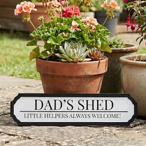 Puede incluir: Un letrero en blanco y negro que dice "DAD'S SHED" con el texto "LITTLE HELPERS ALWAYS WELCOME!" debajo. El letrero está frente a una maceta de terracota con flores y suculentas. Otras macetas están en el fondo.
