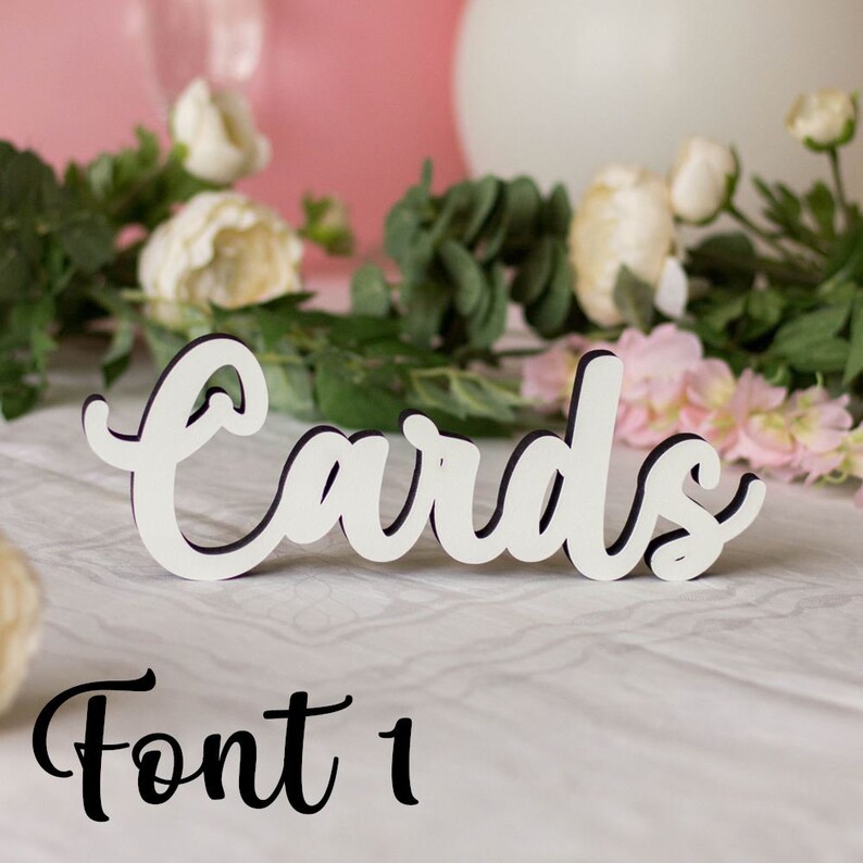 May include: White wooden sign with the word "Cards" written in a cursive font. The sign is sitting on a white surface with flowers in the background. The text "Font 1" is written below the sign.