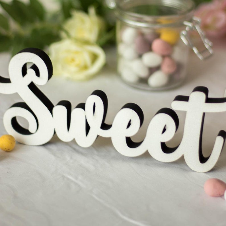 Puede incluir: Un letrero de madera blanco con la palabra "Sweet" en letras negras. El letrero est&aacute; sobre una superficie blanca con un tarro de caramelos en el fondo.
