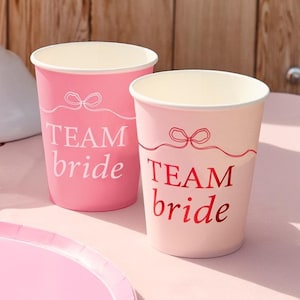 May include: Two pink paper cups with the text "TEAM bride" printed on them. The cups have a white interior and a bow design above the text. One cup is a darker pink than the other.