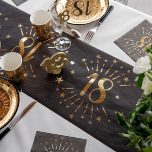 Puede incluir: Un camino de mesa negro y dorado con un diseño de '18' dorado y estrellas doradas. El camino de mesa está sobre un mantel blanco con artículos de fiesta dorados y negros.