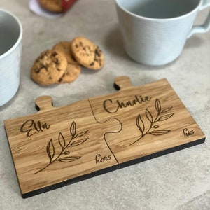May include: Two wooden puzzle piece coasters with the names "Ella" and "Charlie" engraved on them. The coasters are joined together and have a small branch design with leaves and berries. The words "hers" and "his" are also engraved on the coasters.