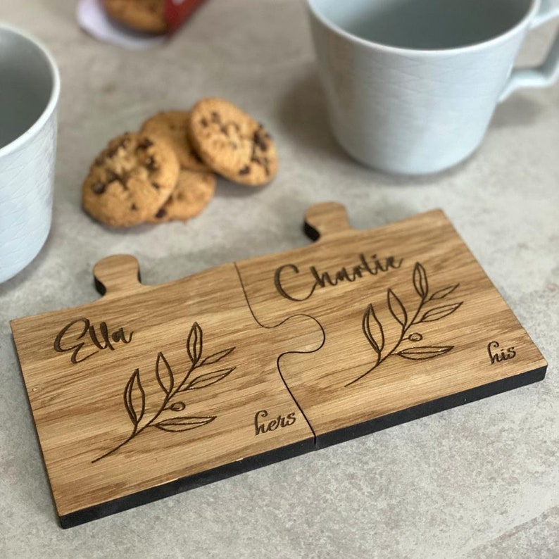 May include: Two wooden puzzle piece coasters with the names "Ella" and "Charlie" engraved on them. The coasters are joined together and have a small branch design with leaves and berries. The words "hers" and "his" are also engraved on the coasters.