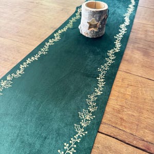 Op de afbeelding: Een donkergroene fluwelen tafelloper met een gouden hulstblad- en bessenontwerp langs de randen. Een kleine houten kandelaar met een stervormige uitsparing staat op de loper, die op een houten oppervlak ligt.