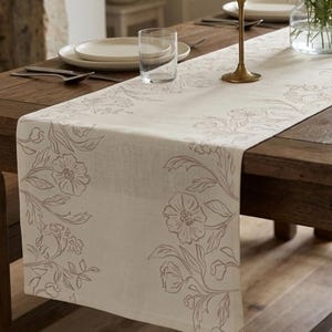 Può includere: Runner da tavolo beige con un motivo floreale beige più scuro. Posizionato su un tavolo di legno scuro, con piatti, posate e un bicchiere. Il motivo floreale è concentrato sui bordi.