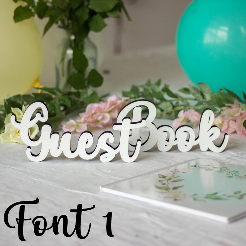 May include: White wooden sign with the words "Guest Book" in a cursive font. The sign is sitting on a white table with a white book with a floral design on the cover.
