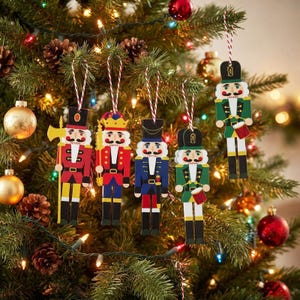 May include: Five flat wooden nutcracker ornaments hanging from red and white striped strings. Each nutcracker features a unique uniform in red, blue, or green, with painted details including hats, mustaches, and accessories like axes or drums.