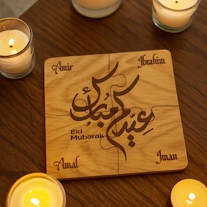 May include: A wooden puzzle with the words "Eid Mubarak" and names "Amir", "Ibrahim", "Amal", and "Iman" engraved on it. The puzzle is made of light-colored wood and is surrounded by lit candles on a dark wooden surface.