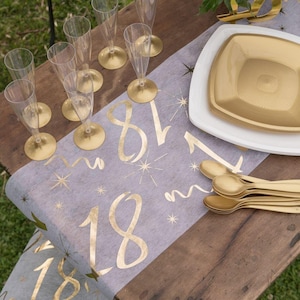 Peut inclure: Un chemin de table doré avec le numéro 18 imprimé en écriture dorée. Le chemin de table est décoré d'étoiles dorées et est posé sur une table en bois. Il y a des assiettes en plastique dorées et des cuillères en plastique dorées sur la table.
