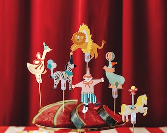 May include: A decorative arrangement featuring watermelon slices on a gold-toned pedestal, adorned with whimsical circus-themed paper cutouts. The cutouts include a lion, zebra, clown, and carousel horse, set against a red curtain backdrop.