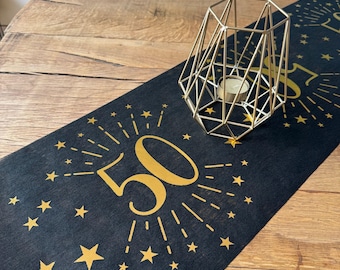 May include: Black table runner with gold foil stars and the number 50. The number 50 is surrounded by gold foil rays. A gold metal candle holder is on top of the runner.