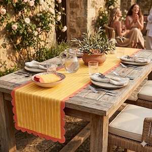 May include: A wooden table set for dining outdoors, featuring a yellow and white striped table runner with a scalloped coral edge. The table is set with plates, silverware, glasses, and a pitcher. A terracotta pot with greenery and dried citrus sits in the center.