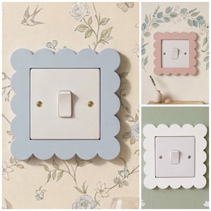 May include: Three decorative light switch covers in pastel shades: light blue, pink, and white. Each has a scalloped edge and surrounds a white light switch. The switches are mounted on walls with floral wallpaper.