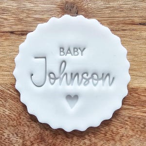 Könnte beinhalten: Ein weißer Stempel mit gezacktem Rand mit den Worten "BABY Johnson" und einem Herzsymbol. Der Stempel ist in eine weiße Oberfläche gepresst, und der Hintergrund ist eine Holzoberfläche.