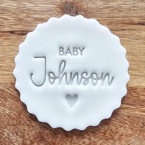 May include: A white, scalloped-edge stamp with the words "BABY Johnson" and a heart symbol. The stamp is made of a solid white material and is set against a wooden background.