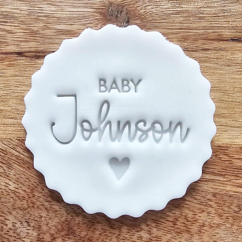 May include: A white, scalloped-edge stamp with the words "BABY Johnson" and a heart symbol. The stamp is made of a solid white material and is set against a wooden background.