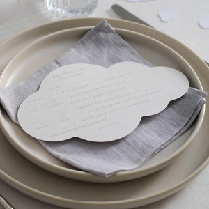 Peut inclure: Une carte de prédiction de baby shower en forme de nuage, avec des sections à remplir pour le nom, la couleur des cheveux et des yeux, posée sur une serviette en lin grise et des assiettes beiges. La carte propose des questions sur l'apparence du bébé.