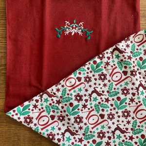 May include: Red fabric with a white and green embroidered design. The fabric is folded to reveal a white fabric with a repeating pattern of green leaves, red hearts, red stars, and the word "JOY" in red.