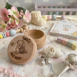 Puede incluir: Una caja redonda de madera grabada con un oso, una mariposa y el texto Cara's FIRST TOOTH. Incluye un dije de zapato de bebé color plata en una cadena, un adorno blanco en forma de nube con un diente, un libro CARA'S FIRSTS y bloques de colores pastel.