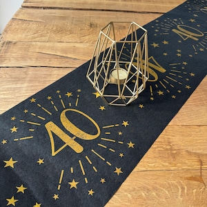 May include: A black table runner with gold accents, featuring the number "40" in a celebratory design with starbursts and stars. A gold geometric candle holder with a lit candle sits on the runner. The runner is on a wooden table.