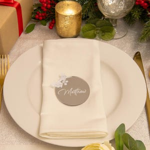 Puede incluir: Una tarjeta de lugar redonda de color plateado con el nombre "Matthew" en escritura cursiva, adornada con un diseño floral. La tarjeta descansa sobre una servilleta blanca, colocada sobre un plato blanco, con cubiertos dorados y decoraciones festivas en el fondo.
