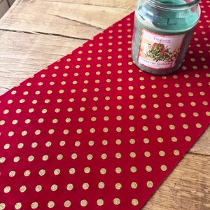 May include: Red table runner with gold polka dots. The table runner is on a wooden table with a glass jar candle that says "Fragrance Under the Mistletoe".