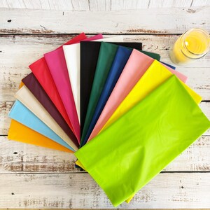 Puede incluir: Abanico de hojas de papel de seda de colores brillantes, incluyendo rosa, verde, amarillo y azul, sobre una superficie de madera clara. Se observa un tarro con líquido amarillo en el fondo.