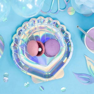 May include: A seashell-shaped iridescent party plate holds two macarons, one pink and one purple, on a light blue surface. The plate is surrounded by party decorations, including balloons and confetti, creating a festive scene.