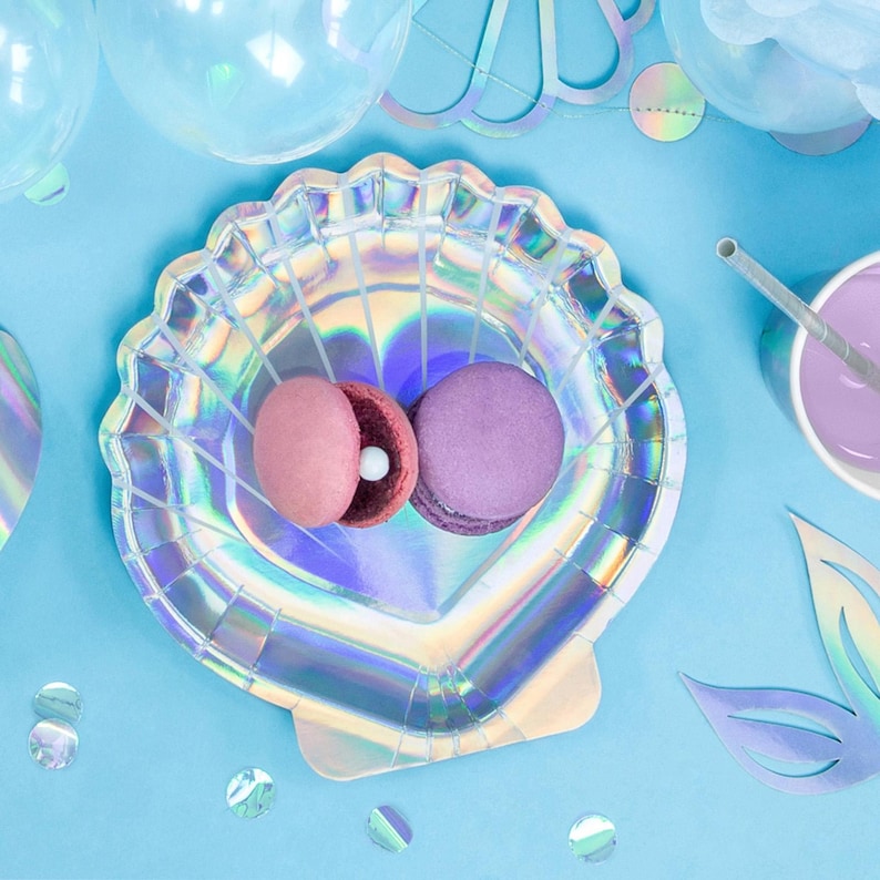 May include: A seashell-shaped iridescent party plate holds two macarons, one pink and one purple, on a light blue surface. The plate is surrounded by party decorations, including balloons and confetti, creating a festive scene.