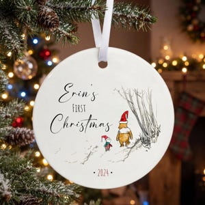Op de afbeelding: Een rond wit keramieken ornament hangend aan een wit lint. Het ornament bevat de zwarte tekst Erin's First Christmas, een kleurrijke illustratie van Winnie de Poeh en Knorretje met kerstmutsen, en het jaartal 2024 in rode letters.