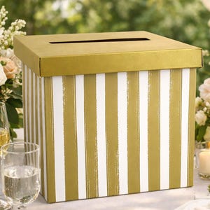 Könnte beinhalten: Eine rechteckige Kartenbox mit einem metallisch goldenen Deckel und einem Körper, der mit vertikalen weißen und goldenen Streifen verziert ist. Der Deckel enthält einen horizontalen Schlitz in der Mitte der Oberseite zum Einwerfen.