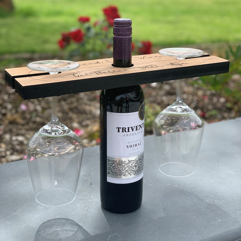 May include: A wooden wine caddy with a bottle of red wine and two wine glasses. The caddy has a hole for the bottle and slots for the glasses. The caddy has the words "Get Better With Age" engraved on it.